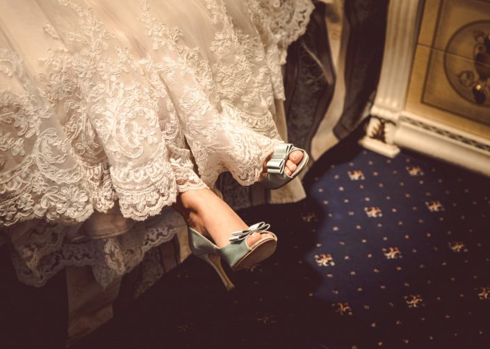 Eine Braut sitzt in ihren Hochzeitskleid auf einem Bett. Sie hat blaue Hochzeitsschuhe an.